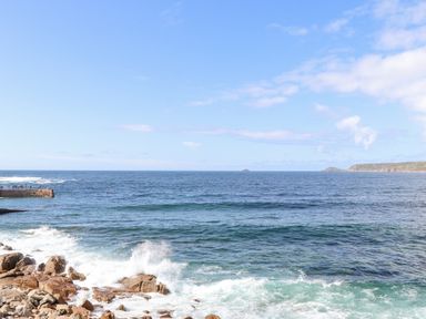 A rocky coastline with waves crashing against rocks and a pier with people at Harbour Studio in Sennen Cove