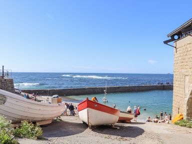 A coastal scene with boats on a ramp leading to the sea and people swimming and walking near a stone pier at Harbour Studio in Sennen Cove
