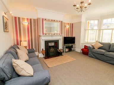 A living room with gray sofas a fireplace a wall mirror a television on a glass stand and a window with blinds at Westholme Lodge in Minehead