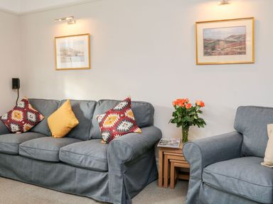 A living room with a sofa and armchair with cushions a side table with flowers and magazines and framed pictures on the wall at Westholme Lodge in Minehead