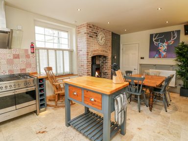 A kitchen with a wooden island unit a dining table with chairs a rocking chair a brick fireplace and a colorful deer painting at Westholme Lodge in Minehead