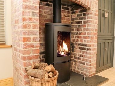 A wood burning stove with fire inside set in a brick fireplace with a basket of logs beside it at Westholme Lodge in Minehead