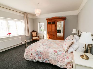 A bedroom with a double bed with floral bedding a wooden wardrobe an armchair bedside tables with lamps and windows with curtains at Westholme Lodge in Minehead
