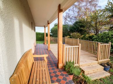 A covered porch with wooden bench and tiled floor next to a wooden deck with railings and steps surrounded by plants