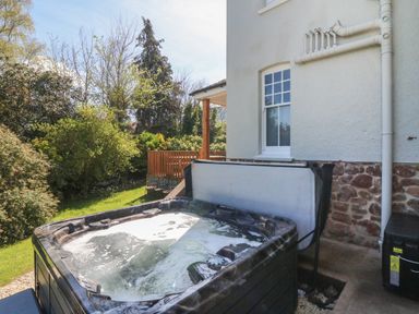An outdoor hot tub next to a house with a window and a garden with trees and shrubs at Westholme Lodge in Minehead