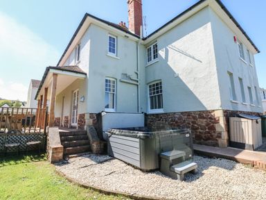 The back exterior of a two story house with a small covered porch stairs leading to the door a hot tub on a gravel area and a grassy lawn at Westholme Lodge in Minehead