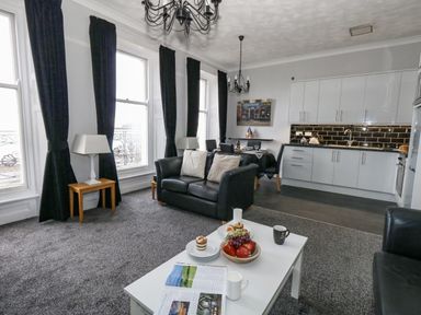 An open plan living room and kitchen with black leather sofas dining table white cabinets and large windows with black curtains at Sunbeach in Weymouth