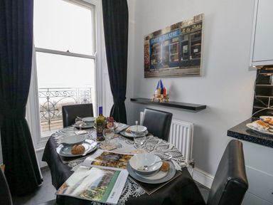 A dining table set for four with bowls plates cutlery a bottle of wine glasses and a magazine near a window with black curtains at Sunbeach in Weymouth