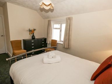 A bedroom with a bed with folded towels and red pillows a black dresser with a vase of flowers and two wicker chairs by window at Sunbeach Weymouth