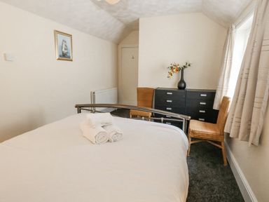 A bedroom with a bed and folded towels on it two wicker chairs a black dresser with a vase of flowers and a window with curtains at Sunbeach in Weymouth