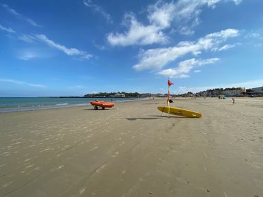 A sandy beach with lifeguard equipment including a yellow surfboard and an orange boat near the shoreline at Sunbeach in Weymouth
