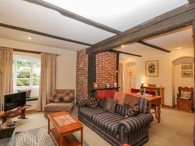 A living room with striped sofa, wooden coffee table, TV, armchair, dining table, exposed wooden beams, and brick column at Field Cottage in Shepton Mallet