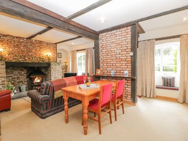 A living room with a wooden dining table and chairs with red cushions a brick fireplace and a sofa at Field Cottage in Shepton Mallet