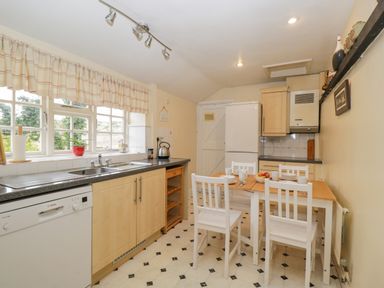 A kitchen with wooden cabinets a dining table with four white chairs and a window with a checked curtain at Field Cottage in Shepton Mallet