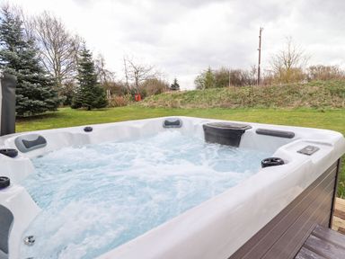 An outdoor hot tub with bubbling water in a grassy area with trees and a hill in the background at Lodge 1 in South Hykeham