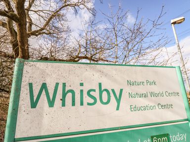 A sign for Whisby Nature Park Natural World Centre and Education Centre with trees and a lamppost in the background