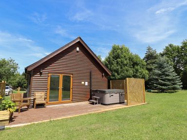 A wooden cabin with a deck patio chairs and a hot tub surrounded by trees at Lodge 1 in South Hykeham