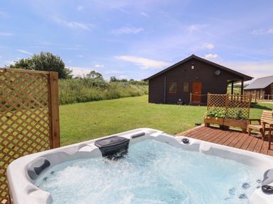 An outdoor hot tub on a wooden deck with a wooden cabin and green lawn in the background at Lodge 1 in South Hykeham