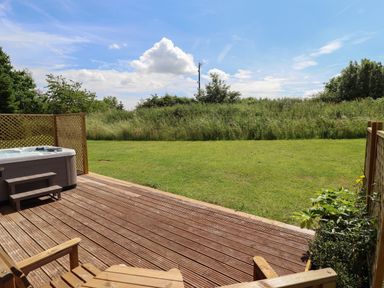 A wooden deck with a hot tub and chairs overlooking a grassy yard and dense greenery at Lodge 1 in South Hykeham