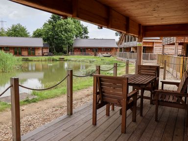 A covered wooden deck with table and chairs overlooking a pond and wooden lodges at Lodge 24 in South Hykeham