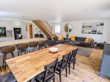 An open plan living area with a wooden dining table and black chairs a kitchen island with bar stools gray sofas with cushions and a staircase in the background at Lodge 24 in South Hykeham