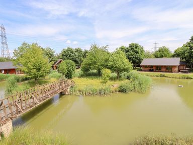 A wooden bridge over a pond with trees and two wooden houses in the background at Lodge 24 South Hykeham