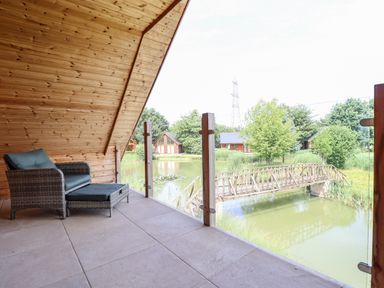 A balcony with a wicker chair and footrest overlooking a pond with a wooden bridge and trees at Lodge 24 in South Hykeham