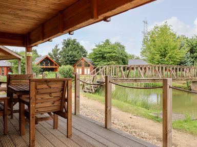 A covered wooden deck with table and chairs overlooking a pond with a wooden footbridge and cabins in the background at Lodge 24 in South Hykeham