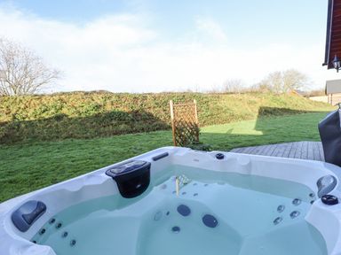 An outdoor hot tub filled with water on a wooden deck with grass and bushes in the background at Lodge 7 in South Hykeham