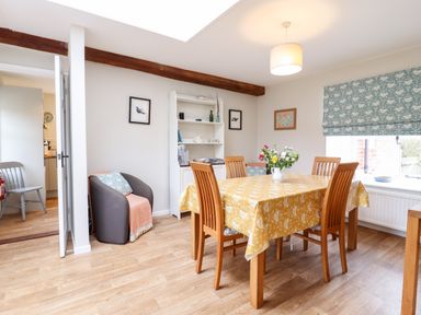 A dining room with a wooden table covered by a yellow tablecloth and four chairs a grey armchair with a cushion and throw a white shelving unit with dishes framed pictures on the walls and a window with patterned blinds at The Brambles in Polstead