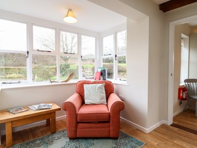An armchair with a cushion next to a wooden bench with magazines in a room with large windows at The Brambles in Polstead
