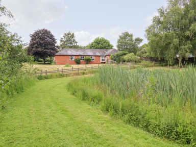A brick house by a wooden fence with a grassy pathway and tall plants near a pond at The Brambles in Polstead