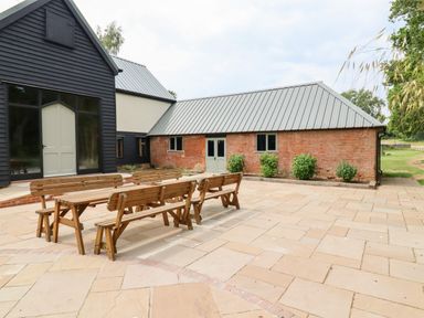 An outdoor patio area with wooden benches and tables in front of buildings with black, white, and brick walls at The Brambles in Polstead