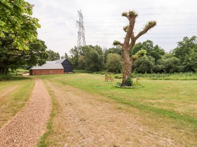 A gravel path leading to a brick and black barn with a grassy area with a tree and benches at The Brambles in Polstead