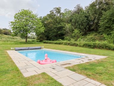 An outdoor rectangular swimming pool with a pink flamingo float and a pool cover on a surrounding stone patio with grass and trees at The Brambles in Polstead