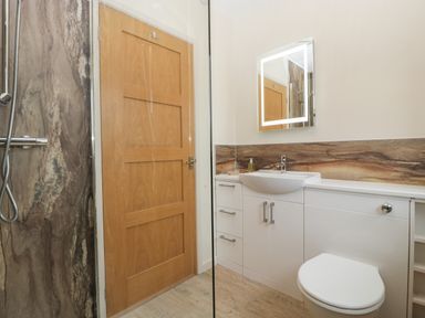 A bathroom with a wooden door a shower with stone wall a white sink with cabinetry and a lighted mirror at Palace Bungalow in St Weonards