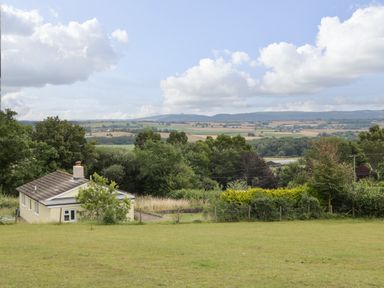 A yellow bungalow with a chimney surrounded by trees and grass with hills and fields in the background at Palace Bungalow in St Weonards