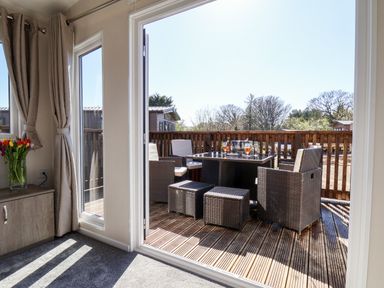 A patio with wicker chairs and a table set with drinks viewed from inside a room with curtains and flowers on a cabinet at Primrose Lodge in Runswick Bay near Staithes