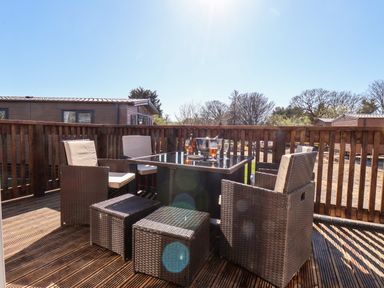 An outdoor wooden deck with a glass table surrounded by wicker chairs and stools with drinks and an ice bucket on the table at Primrose Lodge in Runswick Bay near Staithes