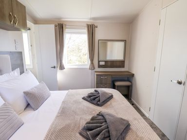 A bedroom with a bed, folded towels on the bed, a window with curtains, a dressing table with stool and a mirror at Primrose Lodge in Runswick Bay near Staithes