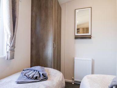 A bedroom with two single beds a wooden wardrobe a wall mirror and a window with curtains at Primrose Lodge in Runswick Bay near Staithes