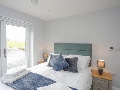 A bedroom with a bed with blue and gray pillows bedside tables with lamps and a glass door to the outside at Tan Y Coed - Nasareth Nasareth near Penygroes