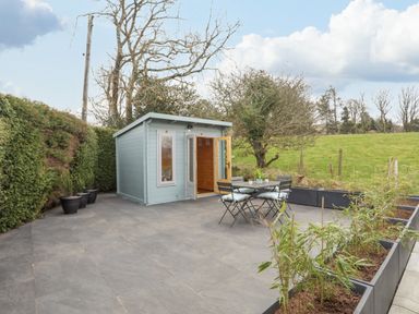 A small blue painted wooden shed with open doors and a table with four chairs on a tiled patio with plants in pots and planters at Tan Y Coed - Nasareth Nasareth near Penygroes