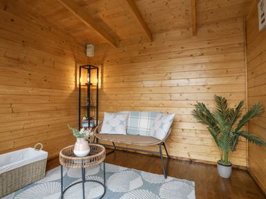 A small wooden room with a wicker bench with pillows a round wicker table with a vase a tall black shelf with a lamp books a plant in a pot and a woven basket on a rug at Tan Y Coed - Nasareth Nasareth near Penygroes