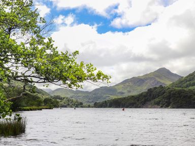 A lake with trees and mountains in the background and a branch with leaves in the foreground at Tan Y Coed - Nasareth Nasareth near Penygroes