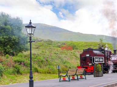 A train station platform with benches a lamppost and a steam train with hills in the background at Rhyd Ddu