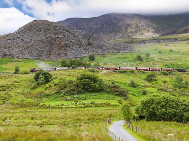 A steam train traveling through a green countryside with hills and a gravel path at Tan Y Coed - Nasareth near Penygroes