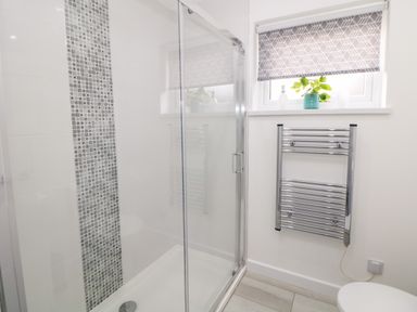 A bathroom with a glass shower enclosure a tiled wall with a mosaic feature a chrome towel radiator a window with a patterned blind and a small plant on the window sill at Harry's Lodge in Ashover