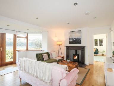 A living room with a pink sofa, green armchair, wooden chest coffee table, fireplace, and large windows at Ynys Faelog Cottage in Ynys Faelog near Menai Bridge