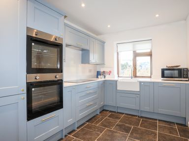 A kitchen with blue cabinets a double oven a microwave a white sink under a window and brown tiled floor at Ynys Faelog Cottage in Ynys Faelog near Menai Bridge